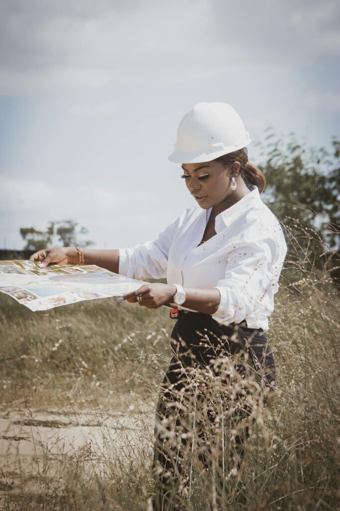 A female engineer wearing a safety helmet reviews construction plans in a grassy field, under a bright sky.